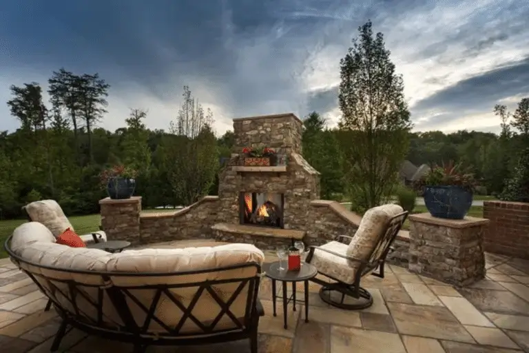 A cozy stone patio by Hearth & Home Specialties features cushioned seating around an outdoor fireplace, nestled in a lush green yard beneath a dramatic dusk sky. Potted plants and small tables create a welcoming touch.