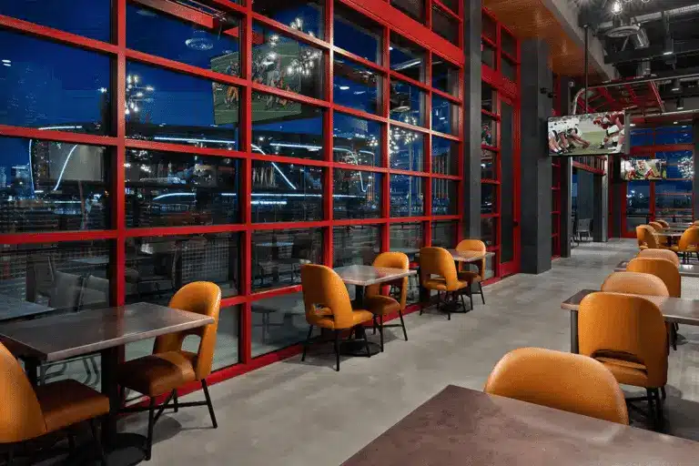 Modern restaurant interior with orange chairs and dark tables, featuring Hearth & Home Specialties. Floor-to-ceiling red-framed windows reveal a nighttime city view, while several TVs show sports in the well-lit space.