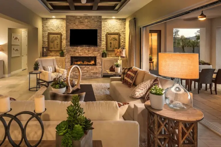 A cozy living room with beige sofas, a stone fireplace by Hearth & Home Specialties beneath a wall-mounted TV, warm lighting, large windows, potted plants, and an open dining area visible through sliding glass doors.